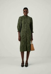 Alyvuogių žalios spalvos midi suknelė su sagų užsegimu, pūstomis rankovėmis ir akių raštu. Papildyta oranžiniais akiniais nuo saulės ir maža ruda rankine.