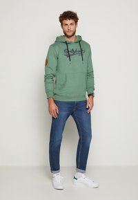 Grüne Kapuzenjacke mit einer Fronttasche, Kordeln und einem Logo-Print. Kombiniert mit körperbetonten blauen Jeans und weißen Sneakern mit grünen Akzenten.