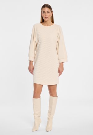 Vrouw draagt een crèmekleurige gewatteerde jurk tot de knieën met lange mouwen en bijpassende knielange laarzen met hakken, staand tegen een witte achtergrond.