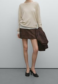 Beżowy sweter z długim rękawem, brązowa mini spódnica ze zamszu, czarne buty na obcasie, trzymająca brązową kurtkę. Gładkie tekstury i proste linie.