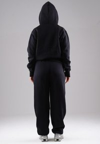 Person standing with back facing camera, wearing black hoodie, black loose pants, and silver athletic shoes against a plain gray background.