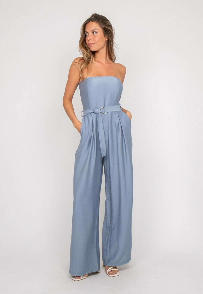 Femme aux longs cheveux portant une combinaison bleu clair à jambes larges sans bretelles avec ceinture et talons blancs ouverts, debout devant un fond uni.