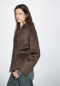 Brown wool blazer with a notched collar, two front pockets, and a relaxed fit, layered over a striped top and paired with blue jeans.