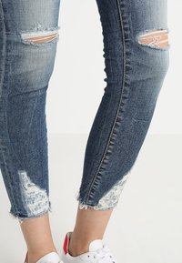 Jeans de mezclilla azul ajustados con detalles desgastados que incluyen rasgaduras y dobladillos deshilachados. Combinados con zapatillas blancas con acentos rojos en el talón.