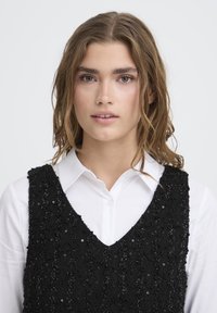 Crni satenasti prsluk s teksturiranim valovitim uzorkom, složen preko bijele košulje sa dugmadima. Izrez je dubokog V-oblik.