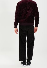 Giacca bomber in velluto bordeaux con grafica triangolare sul retro, abbinata a pantaloni in corduroy neri e sneakers nere.