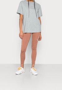Person som bär en ljusgrå Nike-t-shirt, rostfärgade Nike tights med logotyp på låret, och vita Nike-sneakers med oranga snören, står framför en enkel bakgrund.