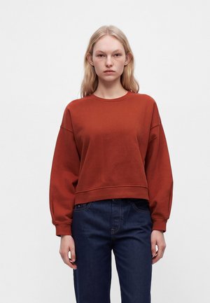 Junge Frau mit glatten blonden Haaren, die einen rostfarbenen Pullover und dunkelblaue Jeans trägt, steht vor einem schlichten weißen Hintergrund.