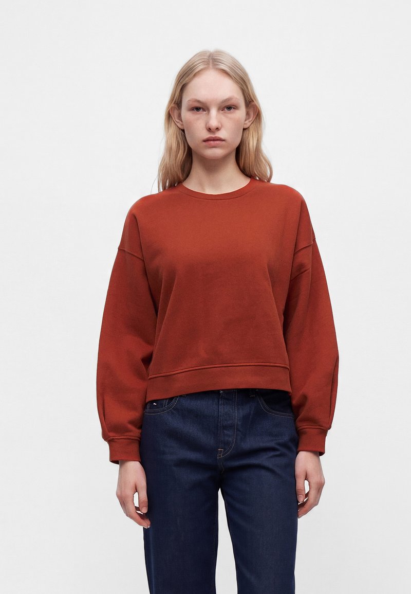 Jeune femme aux cheveux blonds et raides, portant un sweat-shirt couleur rouille et un jean bleu foncé, debout devant un fond blanc uni.