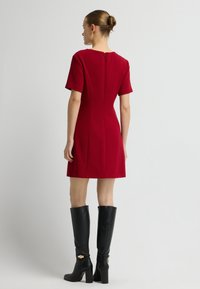 Rode jurk met korte mouwen, een vierkante halslijn en een rits aan de achterkant. In combinatie met kniehoge zwarte leren laarzen met gouden hardware accenten.