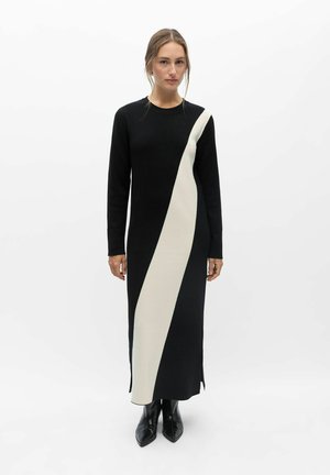 Femme portant une longue robe noire avec une large bande diagonale blanche, debout, les mains détendues et portant des bottes noires pointues.