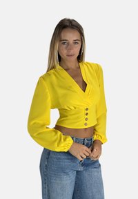 Gul kort crop-top med långa, puffärmar, V-ringning och knappar framtill. Slät textur, kombinerad med högmidjade denimjeans.