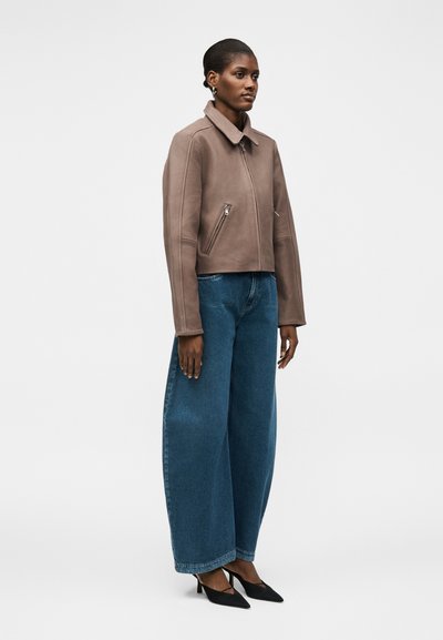 Γυναίκα όρθια, φοράει ένα ταουπ μπουφάν με φερμουάρ, φαρδιά μπλε τζιν και μαύρες μυτερές γόβες, σε απλό λευκό φόντο.