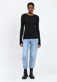 Maglione a maniche lunghe nero a coste con colletto girocollo, abbinato a jeans azzurri con risvolto e stivaletti neri alla caviglia. Dettagli in accentatura d'oro visibili.
