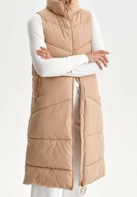 Beige gepolsterte Weste mit hohem Kragen, durchgehendem Reißverschluss und Seitentaschen. Merkmale sind diagonale Steppnähte und eine glatte Textur.