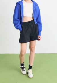 Sudadera azul corta con capucha, top beige corto, pantalones cortos negros y zapatillas bicolor. Las zapatillas tienen cordones negros en un lado y crema en el otro.