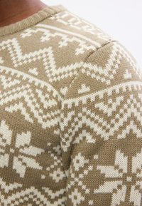 Maglione lavorato a maglia in beige con motivi geometrici e floreali bianchi. Presenta un collo a girocollo a coste e una texture morbida.