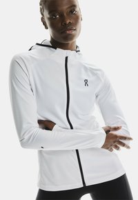 Veste blanche zippée avec un design ajusté, en tissu texturé, fermeture éclair noire et logo. Comprend une capuche et des manches longues avec des trous pour les pouces.