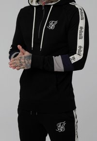 Felpa nera con zip e coulisse bianche, con maniche a blocchi di colore grigio e blu navy, e dettagli con logo sul petto e sulle maniche.