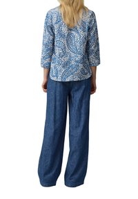 Gemusterte blaue Baumwollbluse mit Dreiviertelärmeln, kombiniert mit lockeren, dunkelblauen Jeans, von hinten vor einem weißen Hintergrund gezeigt.