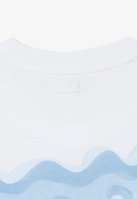 Cuello de camisa blanca con logo de cocodrilo en relieve y patrón abstracto de ondas azules en la tela bajo el cuello.