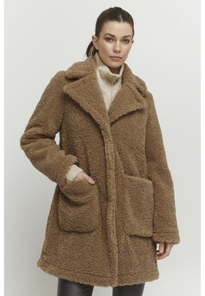 Brown faux fur coat with a notched collar, two front pockets, and a soft texture. Worn over a light sweater, featuring long sleeves.