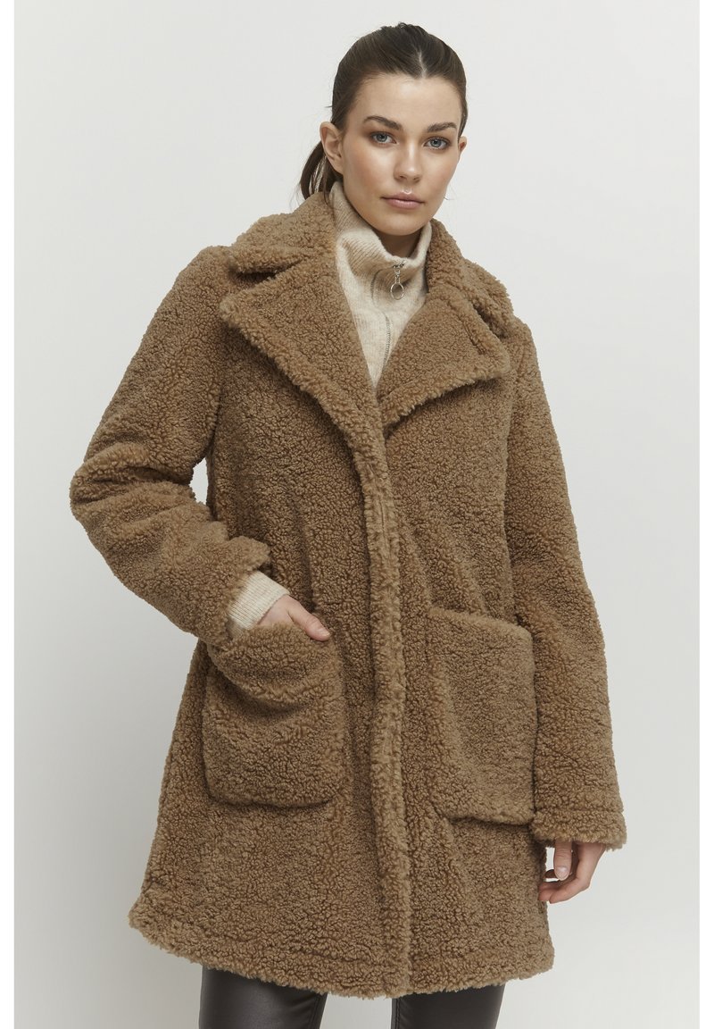 Brun faux fur frakke med en kædekrave, to forlommer og en blød tekstur. Båret over en let sweater med lange ærmer.