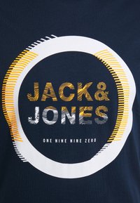 Námořnická modrá bavlněná tričko s kulatým grafickým motivem v bílé a žluté, s textem "JACK & JONES" a "ONE NINE NINE ZERO" pod ním.