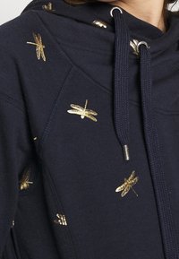 Felpa blu marina con ricamo di libellula dorata, dotata di cappuccio regolabile con cordino e tessuto testurizzato. Il design include piccoli accenti metallici in tutto il capo.