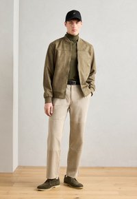 Chaqueta bomber de ante verde oliva, pantalones de algodón beige, zapatos verde oscuro y una gorra negra con logo. El modelo está de pie sobre un suelo de madera.