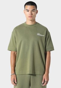 Oliwkowa bawełniana koszulka z luźnym krojem, okrągłym dekoltem i krótkimi rękawami. Posiada małe białe logo na lewej piersi.