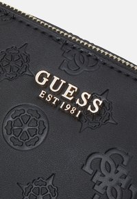 Czarny skórzany portfel z tłoczonymi wzorami kwiatowymi i monogramem, z złotym logo "GUESS EST 1981" oraz złotym zamkiem błyskawicznym.