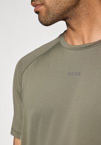 Oliwkowo zielona sportowa koszulka z krótkim rękawem, o okrągłym dekolcie, gładkiej teksturze i logo "BOSS" nadrukowanym ciemniejszym odcieniem na piersi.