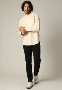 Lichtbeige overhemd met lange mouwen en een mandarinnekraag, knopen aan de voorkant, gecombineerd met zwarte broek en witte sneakers met grijze accenten.