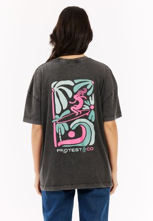 Persoon met lang donker haar die een zwart oversized T-shirt draagt met een kleurrijk abstract ontwerp van surfen en palmbomen op de achterkant.