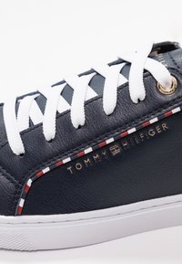 Sneaker in pelle blu navy con lacci bianchi, caratterizzata da un accento a strisce rosse e bianche e dal logo dorato "Tommy Hilfiger" sul lato.