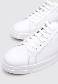 Zapatillas de cuero blancas con un acabado texturizado, cordones planos blancos, diseño de caña baja y suela de goma. Estética limpia y minimalista.