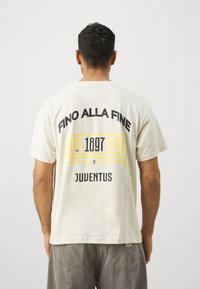 Krémové tričko s čiernym a žltým textom a grafikou na chrbte, vrátane "FINO ALLA FINE," "1897," a "JUVENTUS." Krátke rukávy a štandardný strih.