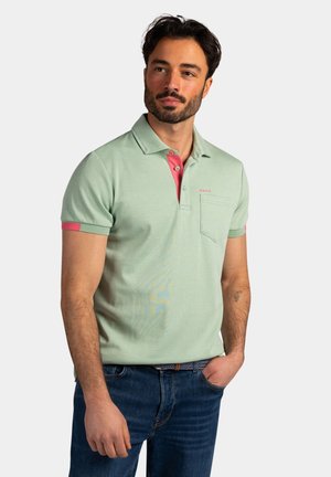 Man met baard, gekleed in een lichtgroen poloshirt met roze kraagdetail en een blauwe spijkerbroek, staand met een hand in de zak, kijkt iets naar rechts.