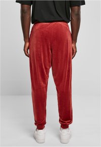 Red velour joggers with cuffed ankles, elastic waistband, and smooth texture, paired with white sneakers, viewed from the back.