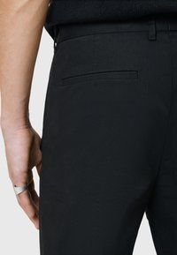 Zwarte op maat gemaakte trousers met een gladde textuur, voorzien van een achterzak en een slank pasvorm ontwerp. Metalen accent ring zichtbaar aan de hand.