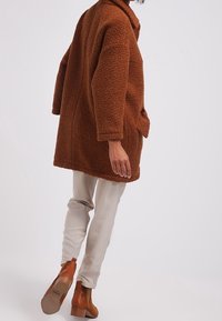 Brun, oversize, strukturerad fleecejacka med krage och framficka, kombinerad med ljusa byxor och beige ankelboots.