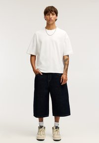 Bílý oversized tričko, tmavé džíny široké šortky, béžové tenisky. Jednoduchý design, pohodlný střih, krátké rukávy, viditelný kovový náhrdelník.