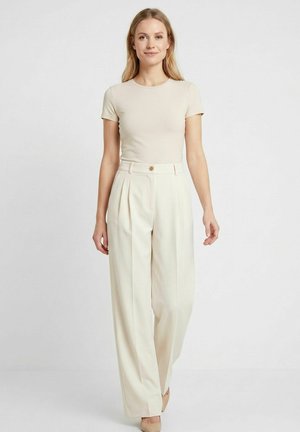 Donna bionda in piedi, indossa un top beige aderente a maniche corte e pantaloni crema a vita alta e gamba larga con dettaglio di bottoni.
