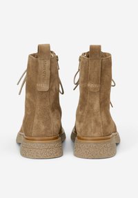 Botines de ante marrón con punta redonda, cremalleras traseras y lazos decorativos. Suelas de goma texturizada con un borde marrón claro.