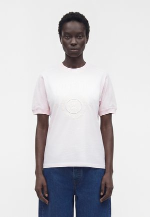 Personne aux cheveux courts et bouclés portant un t-shirt rose clair embossé d'un logo et un pantalon en jean bleu, debout devant un fond blanc uni.