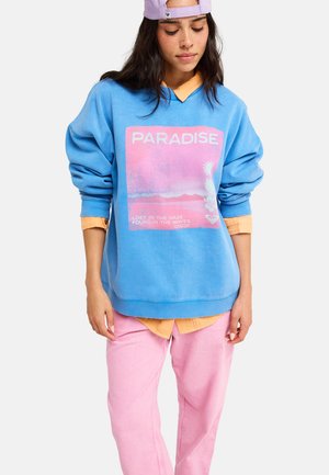 Jonge vrouw die een blauwe trui draagt met de tekst "PARADISE", een oranje shirt eronder, roze broek en een lavendelkleurige pet achterstevoren.