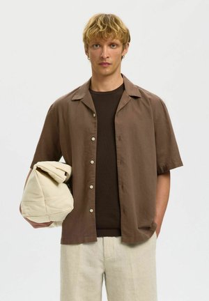 Mladý muž se světle blond krátkými vlasy, oblečený v hnědé krátké košili na knoflíky přes tmavě hnědé tričko, v levé ruce drží složenou světle béžovou bundu.