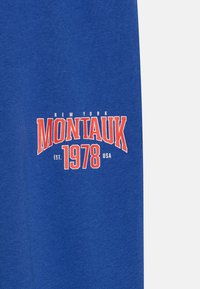 Zils kokvilnas džemperis ar uzrakstu "NEW YORK MONTAUK EST. 1978 USA" sarkanā un baltā krāsā priekšpusē.