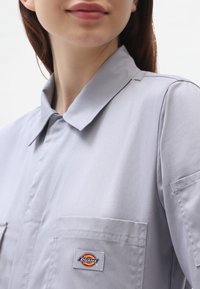 Detailní záběr na osobu v svetle šedé košili s límečkem a přední kapsou, na které se nachází nášivka značky Dickies.